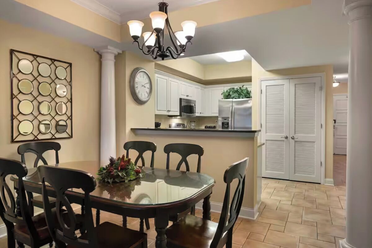Un comedor con mesa de vidrio y sillas de madera, un candelabro, un adorno en la pared y una cocina con armarios y electrodomésticos blancos.