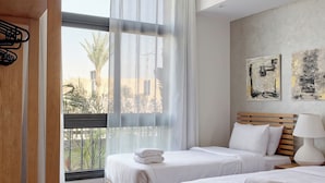 A hotel room with two beds, a wooden wardrobe, a large window with sheer curtains, and artwork on the wall. - Vesta - Luxury Apt - 2BR - CFC (XVI) (New Cairo City)