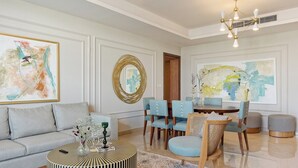 A modern living room with a grey sofa, round coffee table, and blue chairs. There is a dining table with chairs, a large abstract painting, and a chandelier. - Vesta - Luxury Apt - 2BR - CFC (XVI) (New Cairo City)