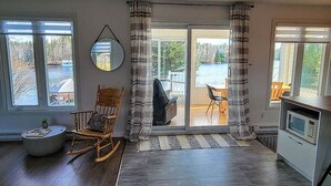 Een ruime woonkamer met een houten schommelstoel, een eettafel met stoelen, een keuken met een magnetron, en grote ramen met uitzicht op een meer en bomen.