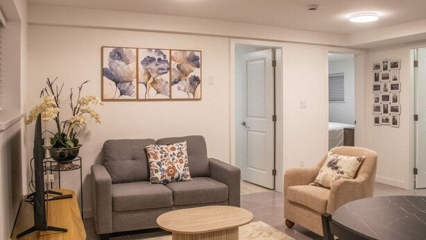 A living room with a grey sofa, a round coffee table, and a beige armchair. There are three wall-mounted paintings above the sofa and a small plant on a stand beside it. - 4615 Inverness Street Vancouver King And Queen 2 Bedrooms Suite Private Entry (Vancouver)