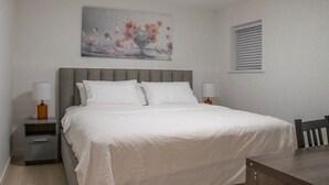 A bedroom with a bed, nightstands, a table, and a window with blinds. - 4615 Inverness Street Vancouver King And Queen 2 Bedrooms Suite Private Entry (Vancouver)