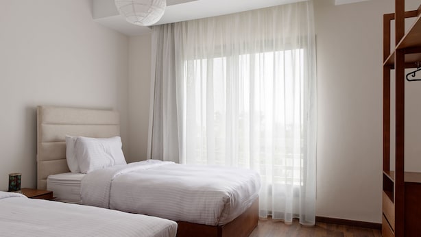 A bedroom with two beds, a wooden headboard, a bedside table, and a large window with sheer curtains. - Vesta - Luxury Apt - 2Br - Cfc (XXXI) (New Cairo City)
