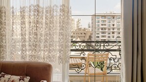 A living room with a brown sofa, floral cushions, sheer curtains, and a view of a balcony with a metal railing and a table. - VESTA - Luxury Apartment - 2BR - Heliopolis (Almazah)