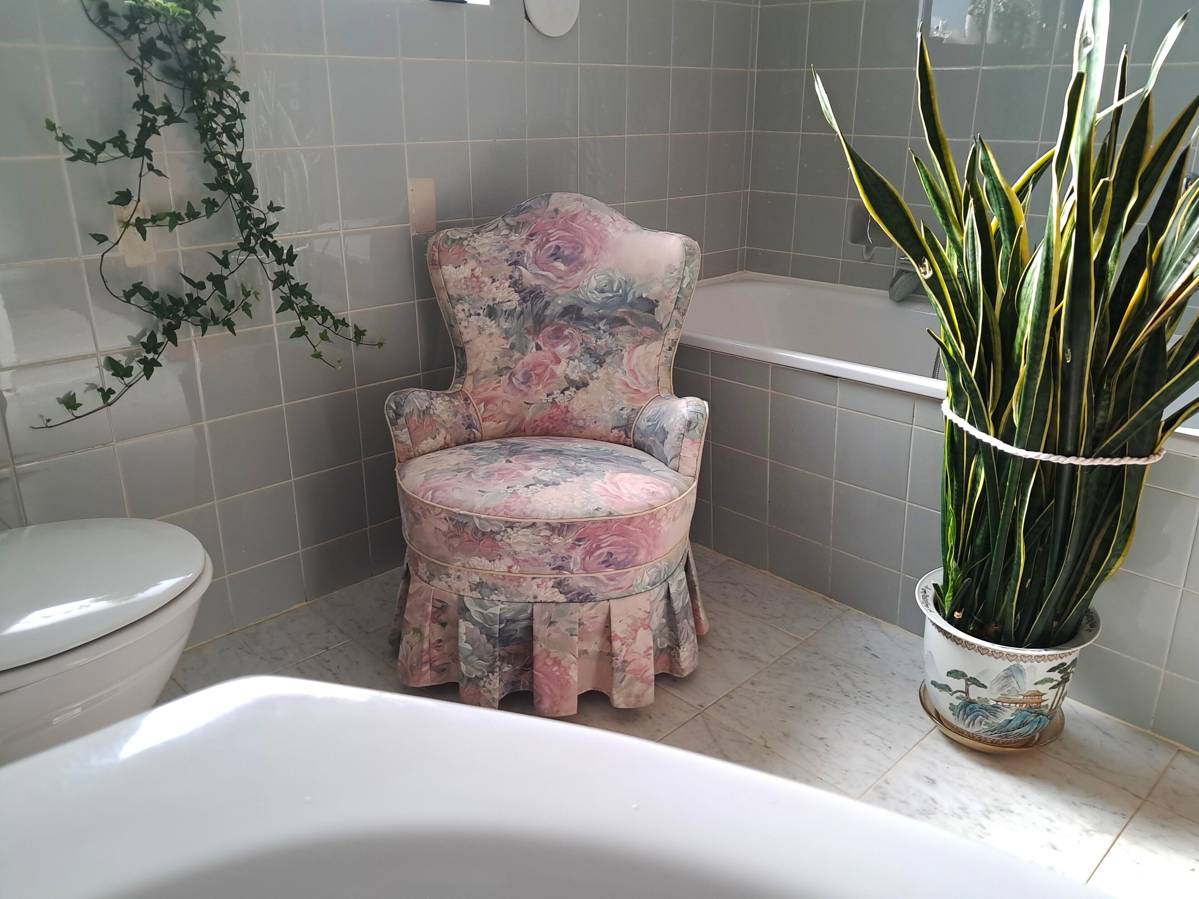 Une salle de bain avec une chaise à motifs floraux, des toilettes, une baignoire et une plante en pot.