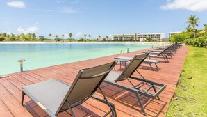 Eine Holzterrasse mit Liegestühlen, die auf ein klares, blaues Meer blickt, mit einem Pool und einem Hotel im Hintergrund.