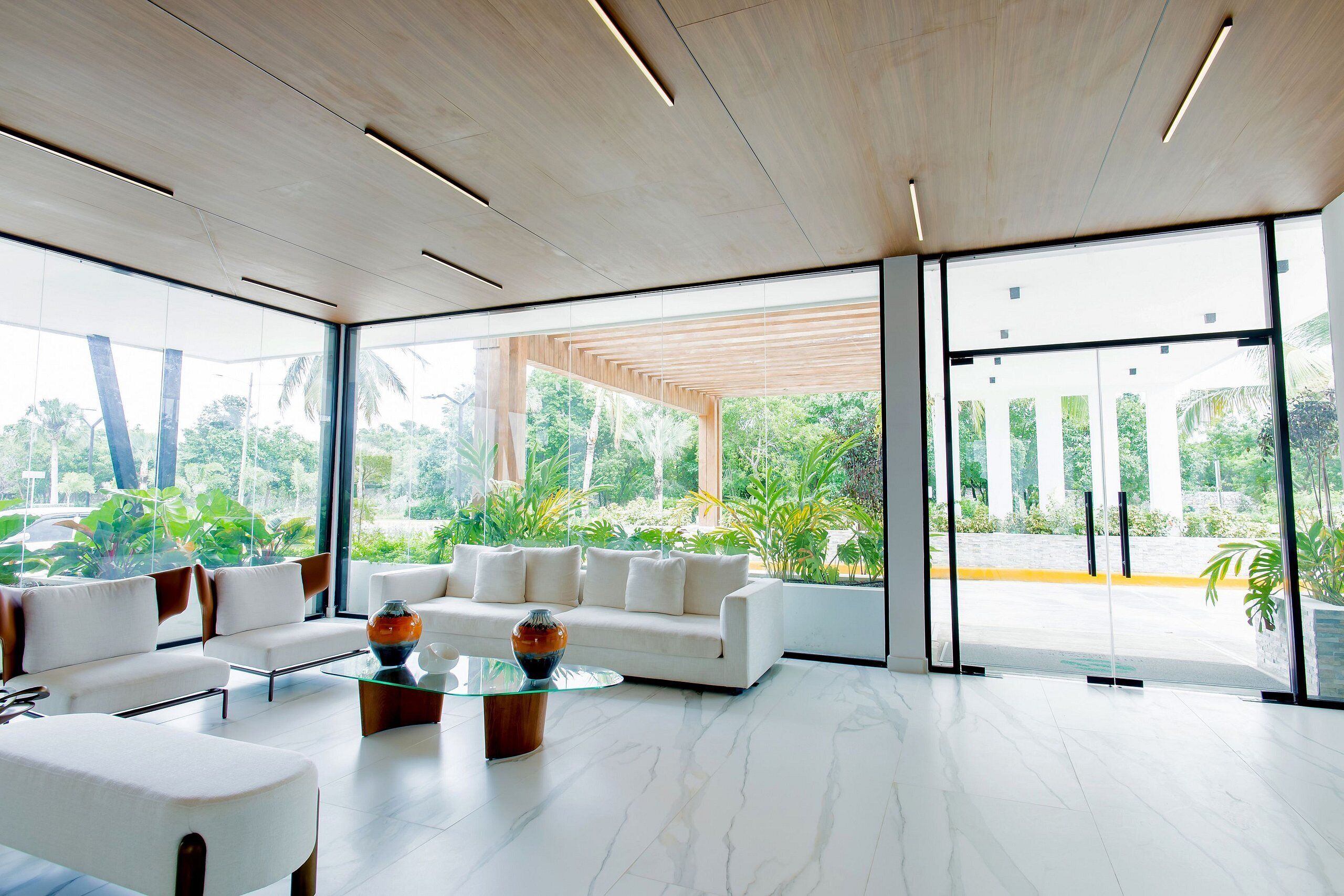 Uma sala de estar moderna com um sofá branco, mesa de centro de vidro e janelas amplas que oferecem vista para o verde lá fora.