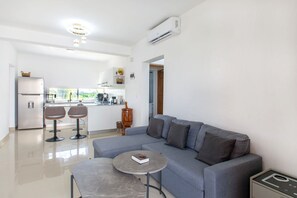 A modern living room with a grey sofa, two bar stools, a coffee table, and a kitchen in the background. - 2 Bedrooms, Pool, 7 Minutes from the Airport (Punta Cana)