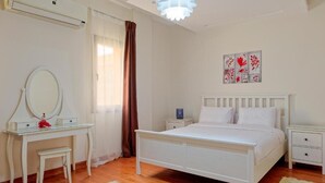 A bedroom with a white bed, a bedside table, a nightstand, a dressing table with a mirror, and a window with curtains.