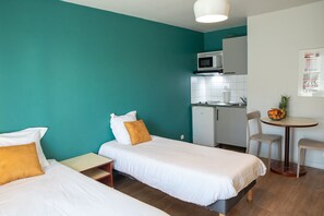 Kompakti hotellihuone, jossa on kaksi erillistä sänkyä, pieni keittonurkkaus ja ruokailutila.