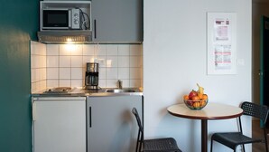 Uma cozinha compacta com micro-ondas, máquina de café e pequena geladeira. Há uma mesa com uma fruteira e duas cadeiras.