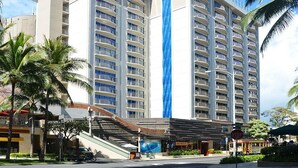 Un hotel de varios pisos con balcones, un elemento vertical azul y palmeras.