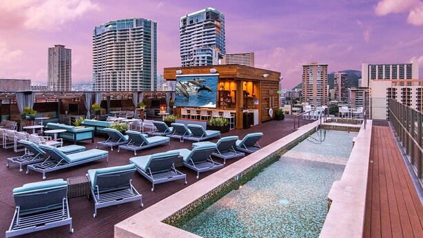 Zona de piscina en la azotea con sillones, bar y vistas a edificios de la ciudad.
