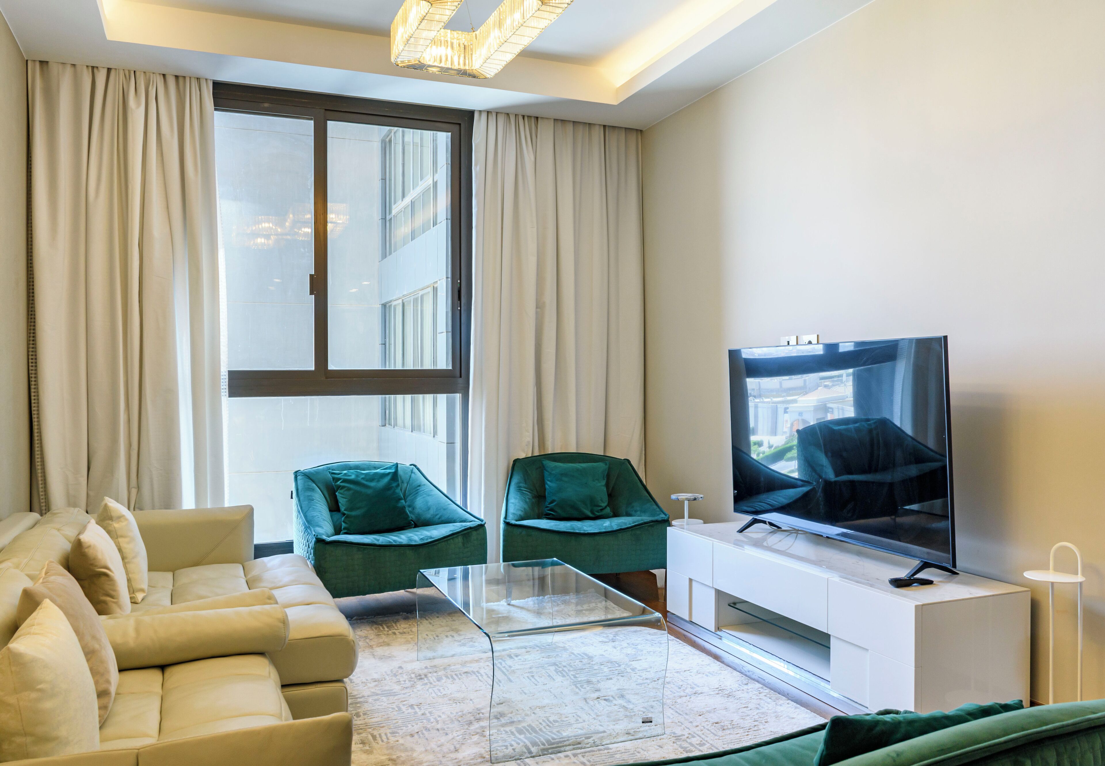 A modern living room with a sofa, two armchairs, a glass coffee table, a flat-screen TV, and a large window with curtains.