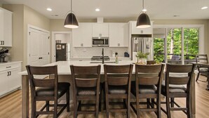 A modern kitchen with white cabinets, a central island, and a dining area with dark wooden chairs. - New Lakefront Estate | Sleeps 34 | Private Dock (LaFollette)