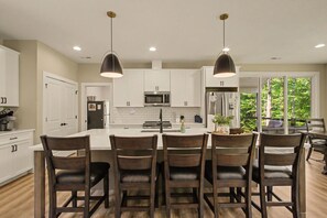 A modern kitchen with white cabinets, a central island, and a dining area with dark wooden chairs. - New Lakefront Estate | Sleeps 34 | Private Dock (LaFollette)
