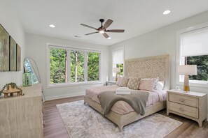 A bedroom with a bed, bedside tables, a nightstand, a ceiling fan, and large windows.