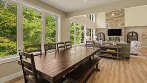 A spacious living room with a large wooden dining table, comfortable seating, a stone fireplace, and large windows offering a view of the greenery outside. - New Lakefront Estate | Sleeps 34 | Private Dock (LaFollette)