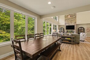 A spacious living room with a large wooden dining table, comfortable seating, a stone fireplace, and large windows offering a view of the greenery outside. - New Lakefront Estate | Sleeps 34 | Private Dock (LaFollette)