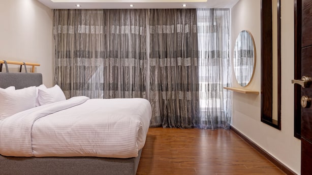 Een slaapkamer met een bed, een houten vloer, een groot raam met gordijnen, een spiegel en een houten deur.
