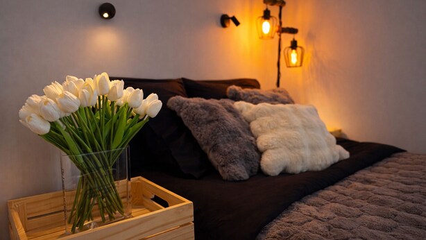 Ein Schlafzimmer mit einem Nachttisch aus Holz, der eine Vase mit weißen Tulpen beherbergt, einem dunklen Bett mit vielen Kissen und Wandleuchten.