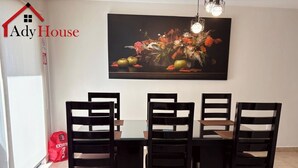 Uma sala de jantar com mesa de vidro e seis cadeiras pretas, um quadro floral grande na parede e uma bolsa vermelha no chão.