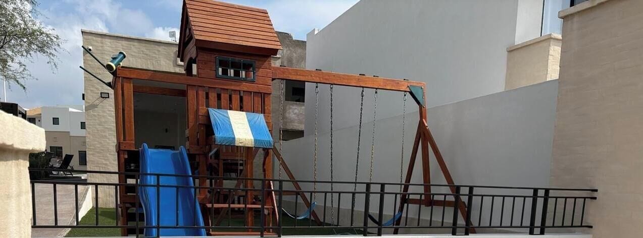 Eine hölzerne Spielanlage mit Rutsche und Schaukeln, umgeben von einem Metallzaun.