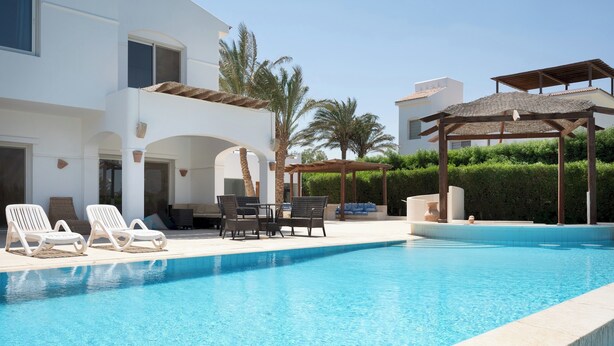 Una piscina con sillas de descanso y una pérgola, rodeada por un edificio blanco con arcos y palmeras.