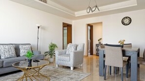 Un salon moderne avec un canapé gris, des coussins à motifs, une table basse ronde, un fauteuil gris, une table à manger avec des chaises et une horloge murale.