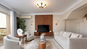 Ein modernes Wohnzimmer mit großem Flachbildfernseher, einer Couch, zwei Sesseln, einem Couchtisch und einer Zimmerpflanze.