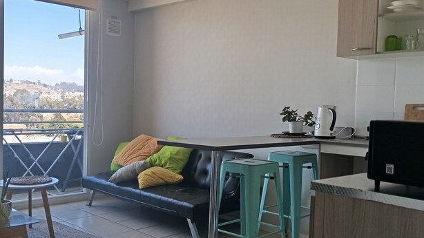 Una cocina moderna con comedor, un sofá con cojines, una mesita y vistas amplias a través de un gran ventanal.
