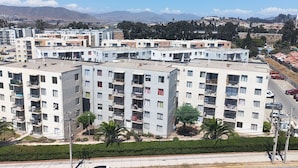 Un edificio residencial de varios pisos con balcones, un área de estacionamiento y una vista despejada de montañas distantes.