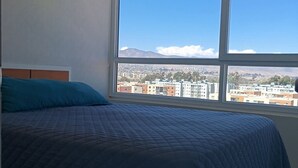 Un dormitorio con un ventanal grande, una cama con colcha azul, y vistas a un paisaje urbano y montañas.