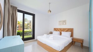 A modern hotel room with a large bed, a bedside table, a nightstand, and a view of palm trees through a large window.