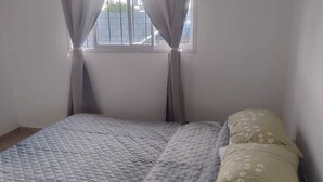 A single bed with a quilted bedspread and floral pillows, a window with curtains, and a plain wall. - Rincon de Olivares, Alta Gracia, Cordoba - Tranquility and Family (Villa del Prado)