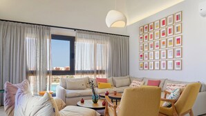 Un salon moderne avec un ensemble de canapés, une table basse et un fauteuil. Les fenêtres sont dotées de rideaux, et un mur est décoré de tableaux encadrés.