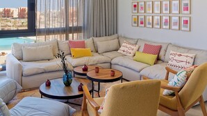 Un salon moderne avec un canapé d’angle, une table basse et deux fauteuils. Il y a une grande fenêtre avec des rideaux, un mur décoré de tableaux encadrés et une vue sur le paysage urbain.