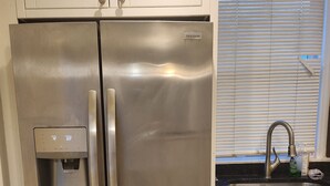 A stainless steel refrigerator with a water dispenser, white cabinets, a kitchen sink, and a window with blinds. - 2-bedroom townhome in superb Baltimore with parking and gated yard (Baltimore)