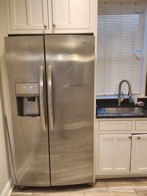 Ein Edelstahl-Kühlschrank mit Wasserspender, weiße Schränke, ein Küchenspüle und ein Fenster mit Jalousien.