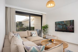 Una sala de estar moderna con un sofá, una mesa de centro con frutas, un televisor y un ventanal con cortinas.