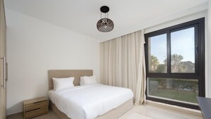 A bedroom with a large bed, a bedside table, a nightstand, a window with curtains, and a view of greenery outside.
