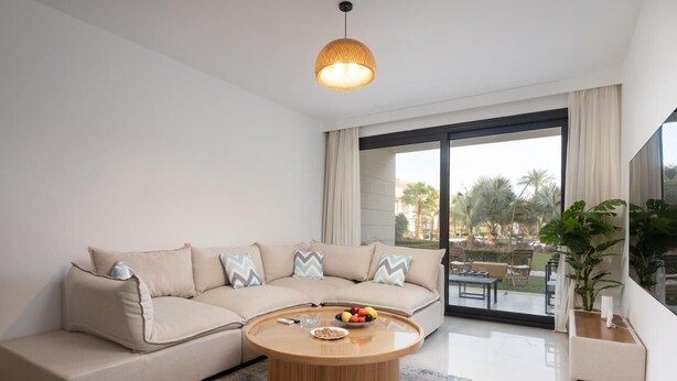 A modern living room with a beige sectional sofa, a wooden coffee table, and a large window with a view of palm trees.
