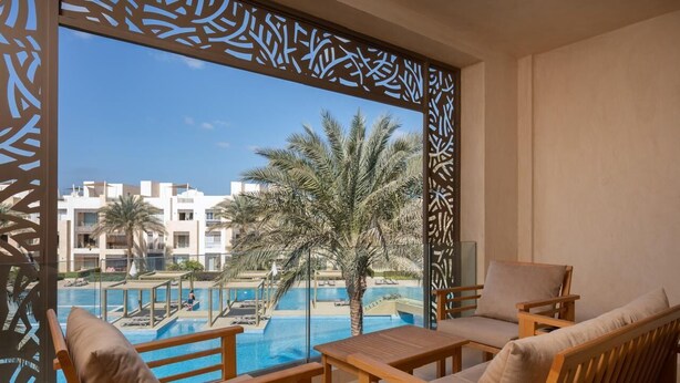 A balcony with wooden furniture, a pool, palm trees, and a view of buildings in the background. - VESTA - Luxury APT - 2BR - Mangrovy (El Gouna)
