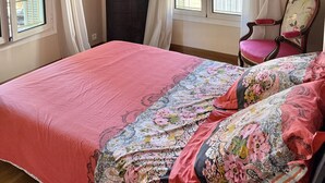 Een slaapkamer met een groot bed, twee ramen met gordijnen, een kroonluchter, een stoel en een dressoir.