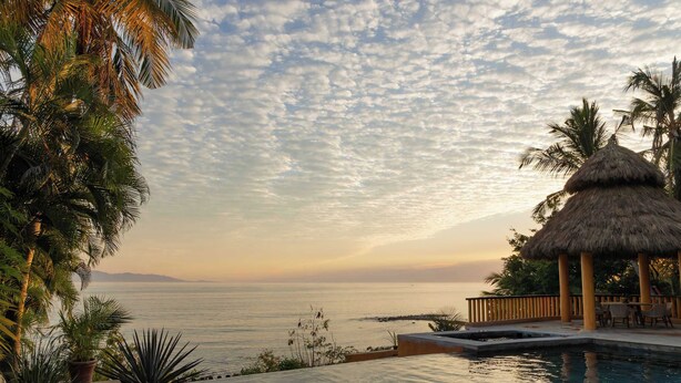 A poolside view with a thatched-roof structure, palm trees, and a clear view of the sea at sunset.