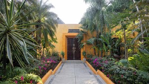 A pathway leading to a building with a dark door, surrounded by lush greenery and flowering plants.