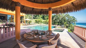 A round table set for dining with wicker chairs, a thatched roof, and a pool in the background.