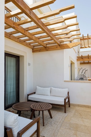 A covered outdoor seating area with a wooden bench and two round tables, a white sofa, and a tiled floor. - VESTA - Luxury APT - 1BR - Fairway (El Gouna)