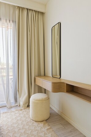 A room with a light-colored wall, a wooden bench, a round ottoman, a mirror, and a window with curtains. - VESTA - Luxury APT - 1BR - Fairway (El Gouna)