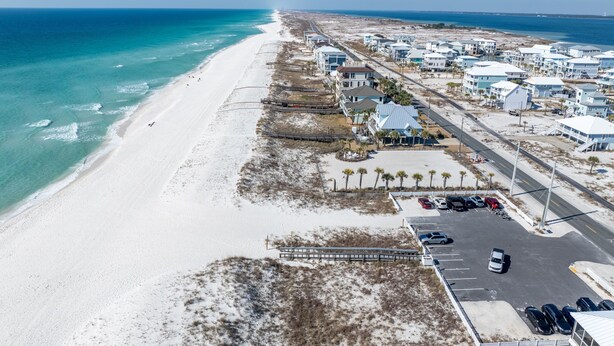 Vista aerea di una zona costiera con spiaggia, una fila di edifici bianchi e un parcheggio con diverse auto.